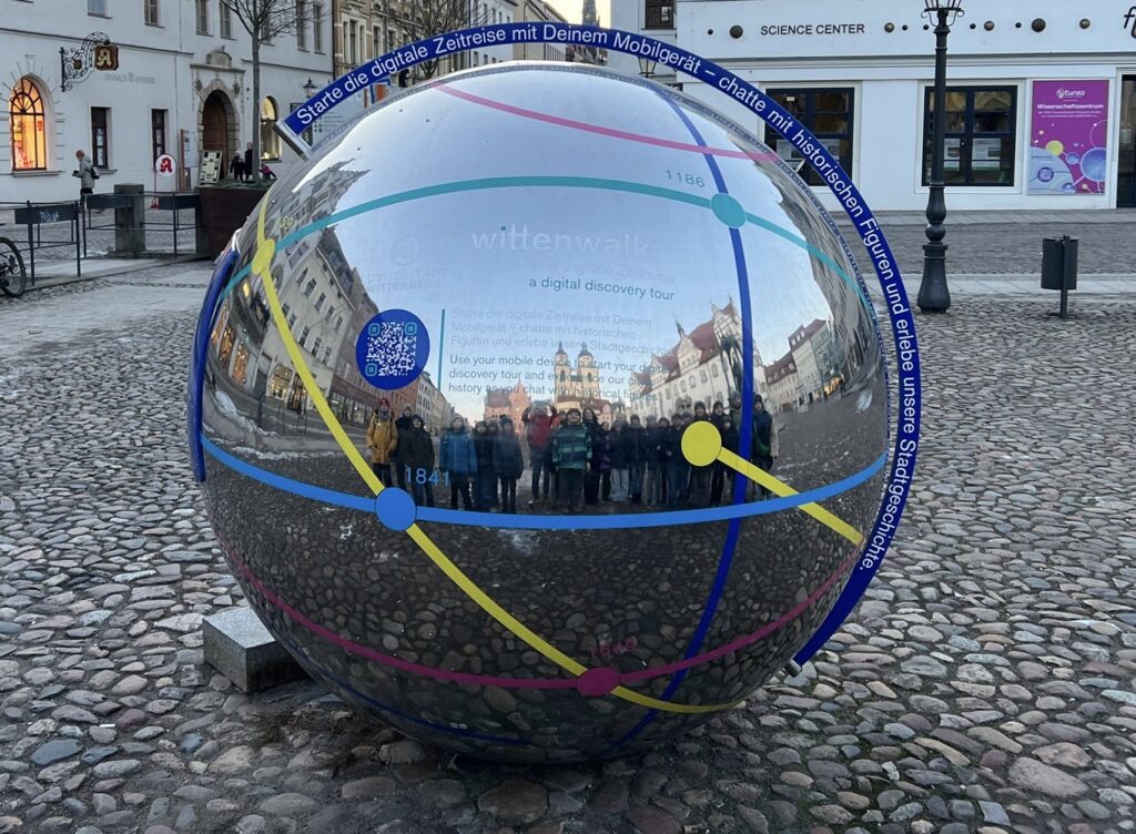 Schüler*innen des Cantor-Gymnasiums während des Spezialistenlagers in Wittenberg, reflektiert in einer runden Metallskulptur mit Zeitreise-Motiv. Im Hintergrund sind die Lutherstadt Wittenberg und der Eingang zum Science Center zu erkennen.