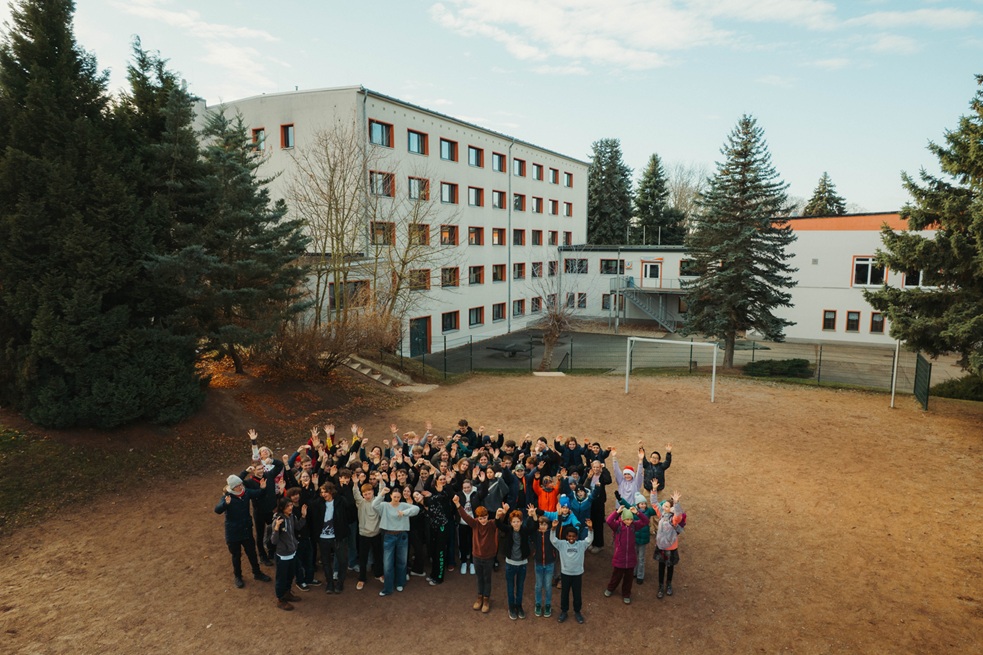 Gruppenfoto der Musik-AGs des Georg-Cantor-Gymnasiums vor der Jugendherberge Naumburg. Die Schülerinnen und Schüler posieren fröhlich auf dem Sportplatz, im Hintergrund sind das Gebäude der Jugendherberge und hohe Bäume zu sehen.
