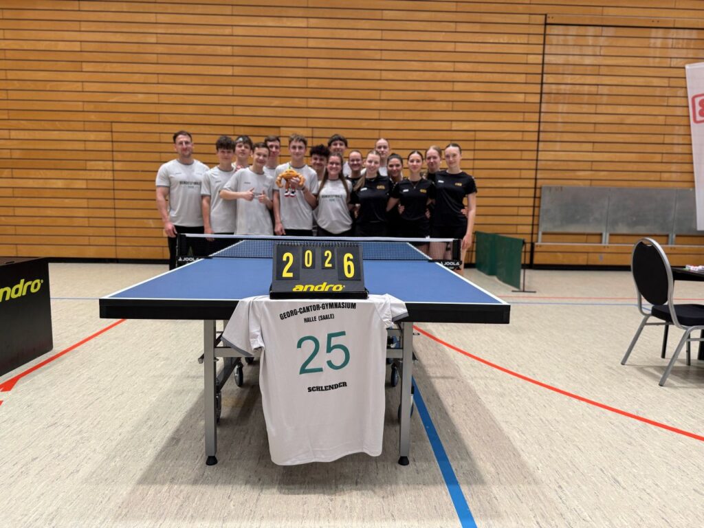Tischtennis-Team des Georg-Cantor-Gymnasiums nach dem Bundesfinale 'Jugend trainiert für Olympia' 2026 in der Sporthalle. Die Mannschaft steht in weißen und schwarzen Trikots hinter einer blauen Tischtennisplatte; darauf liegt ein Trikot mit der Aufschrift 'Georg-Cantor-Gymnasium Halle (Saale)', der Nummer 25 und dem Namen 'Schlender', daneben ein Anzeigeschild mit der Jahreszahl 2026.
